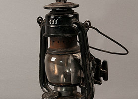 Die Lampe ist aus Metall. Sie hat einen Bügel zum Tragen, und einen Bügel zum Aufgängen. In der Mitte ist ein Glasgefäß. In dem Glasgefäß wird das Feuer entfacht. 