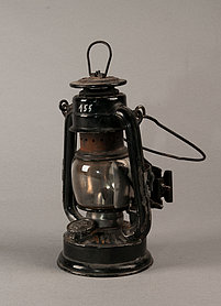 Die Lampe ist aus Metall. Sie hat einen Bügel zum Tragen, und einen Bügel zum Aufgängen. In der Mitte ist ein Glasgefäß. In dem Glasgefäß wird das Feuer entfacht. 