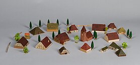 Die Häuser sind klein und aus Holz. Sie haben rote und grüne Dächer. In deer Mitte ist eine Kirche. Manche Häuser sind aus Fachwerk. Manche sind aus Stein. Manche haben sehr große Dächer. Drumherum stehen ein paar Bäume und Zäune.