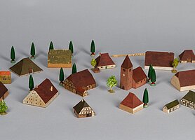 Die Häuser sind klein und aus Holz. Sie haben rote und grüne Dächer. In deer Mitte ist eine Kirche. Manche Häuser sind aus Fachwerk. Manche sind aus Stein. Manche haben sehr große Dächer. Drumherum stehen ein paar Bäume und Zäune.