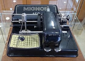 Das Foto zeigt eine schwarze Schreibmaschine. Sie besitzt einen Zeiger, den man zu den Buchstaben und Zeichen führen kann.