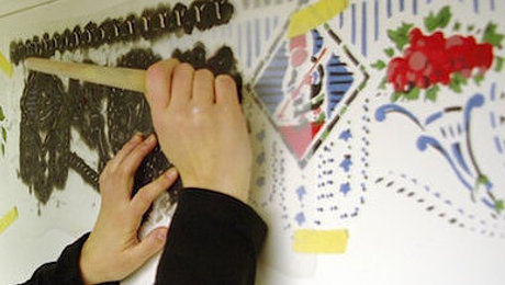 Zwei Hände stupfen mit einem Pinsel durch eine Schablone bunte Muster an eine Wand