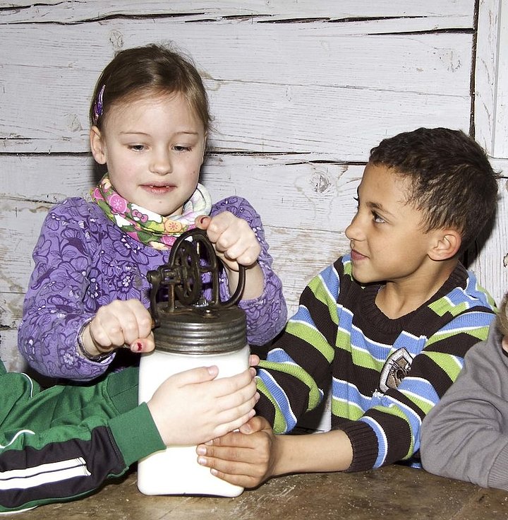Fotoaufnahme der Führung „Von der Kuh zum Butterbrot“.  Um einen Tisch mit einem Butterglas sitzen Kinder. Das Glas wird von zwei Jungen festgehalten, von dem linken Jungen sieht man aber nur die Arme. Das Mädchen in der Mitte dreht an der Kurbel. 
