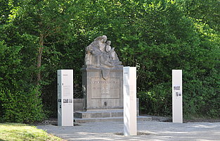 ein Steindenkmal mit drei Figuren - eine "Pietas" mit sterbendem Krieger auf dem Schoß, daneben ein "Jüngling", dem ein Schwert gereicht wird. Drei Informationsstelen kontextualisieren das Denkmal.