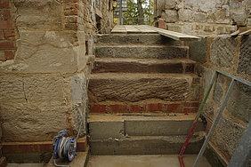 Fotoaufnahme der rekonstruierten Treppe zur Badstube. Die Stufen bestehen aus Sandstein und wurden teilweise mit Ziegelsteinen untermauert. Links befindet sich ein Mauerwerk aus Sandsteinquadern und Ziegeln. Davor steht eine Kabeltrommel, deren Kabel über die Treppe führt. 
