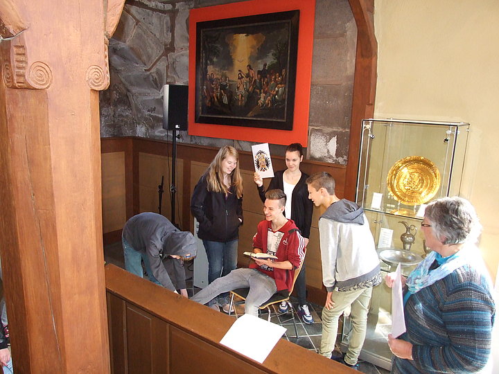 Fotoaufnahme der Aktion "Und woher kommst du?" in der Spitalkirche. Im Fokus steht eine Gruppe aus fünf Jugendlichen und einer Erwachsenen. Mittig sitzt ein Junge mit Buch auf einem Stuhl, das Mädchen dahinter hält ein Bild hoch. Im Hintergrund hängt ein Gemälde der Taufe Jesu. 
