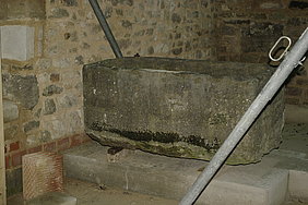 Fotoaufnahme eines Muschelkalkbeckens aus dem Museumsfundus. Das teilweise mit Moos bewachsene Becken befindet sich auf einer Steinplatte, es wurden Holzstücke untergelegt. Im Hintergrund ist ein Bruchsteinmauerwerk erkennbar. 