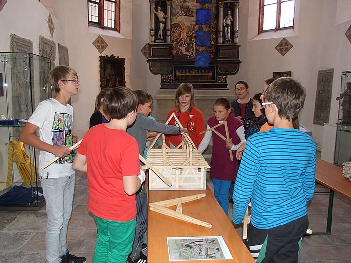 Fotoaufnahme der Führung „Unter dem Dach der Kirche“ in der Bad Windsheimer Spitalkirche. Eine Gruppe Kinder steht um einen Bierzelttisch. Darauf befindet sich ein Haus-Modell, anhand dessen sie ein Dach konstruieren. Im Hintergrund ist ein Altar zwischen zwei Fenstern der Kirche erkennbar.