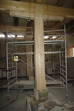 Zwei massive Holzsäulen auf Steinsockel, die Deckenbalken tragen, dazwischen ein Baugerüst. Außerdem Werkzeug und Material zum Befestigen der Säulen.
