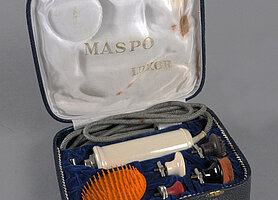 Das Gerät ist länglich und weiß. Es hat ein Kabel. Es hat einen Motor. Es hat verschiedene Aufsätze. Mit den Aufsätzen kann man den Körper massieren. Das ist gesund und soll gut tun. Das Gerät liegt in einem Etui. Das Etui sieht aus wie die Haut einer Schlange.