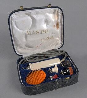 Das Gerät ist länglich und weiß. Es hat ein Kabel. Es hat einen Motor. Es hat verschiedene Aufsätze. Mit den Aufsätzen kann man den Körper massieren. Das ist gesund und soll gut tun. Das Gerät liegt in einem Etui. Das Etui sieht aus wie die Haut einer Schlange.