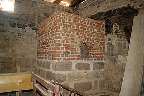 Fotoaufnahme des fertigen Schwitzofens in einer Ecke der Badstube. Der untere Teil besteht aus Sandsteinquadern, der obere wurde mit Ziegelsteinen rekonstruiert. An den beiden sichtbaren Seiten ist je eine rundbogige Öffnung im Ziegelmauerwerk erkennbar. 