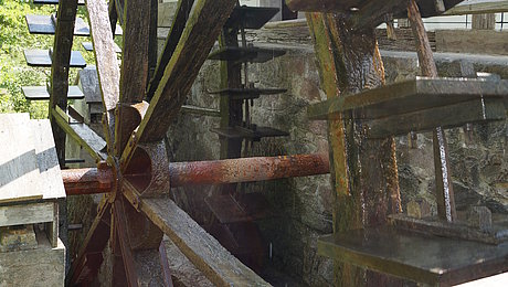 Fotoaufnahme des Wasserrads der Ölmühle von der Flederichsmühle. Im Fokus steht das hölzerne Mühlrad, welches durch ein darunter fließendes Gewässer angetrieben wird. Wasser tropft von dem nassen Rad, das mit Moos bewachsen ist. Dahinter ist die Mühle erkennbar. 