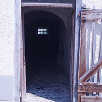 Fotoaufnahme des Eingangs zum Stall aus Reichersdorf am aktuellen Standort. Die Holztür ist um 180° nach außen geöffnet. Hof- und Stallboden sind beide gepflastert, allerding mit unterschiedlichen Pflastersteinen.