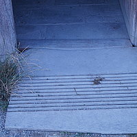 Fotoaufnahme der Schwelle am rechten Eingang der Schäferei aus Hambühl am aktuellen Standort. Vor der Schwelle liegt eine Rampe aus Holz, links und rechts davon wächst Gras. Der Boden im Inneren ist mit Steinplatten gepflastert.
