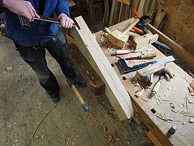 Ein Zimmerer arbeitet mit einem Stemmeisen die Schwalbenschwanzverbindung an einem Kopfholz aus.