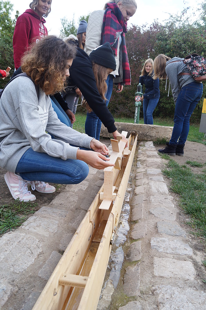 Ein paar Schülerinnen testen ihre selbstgebauten Wasserrädchen in einer Holzrinne an einem Pumpbrunnen