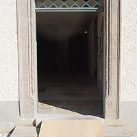Fotoaufnahme des Haupteingangs vom Bauernhaus aus Reichersdorf. Zur Tür führen zwei Steinstufen, über diese wurde eine Rampe aus Holz gelegt. Der Boden des Hofs ist gepflastert, der des Hauses gefliest. 