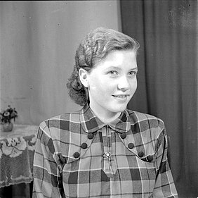 Das Bild zeigt eine Frau. Sie hat eine Frisur mit Wellen. Sie lächelt. Sie trägt eine karierte Bluse.