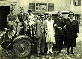 Das Bild zeigt eine Gruppe Frauen und Männer vor einem Auto. Die Menschen sind modisch gekleidet.