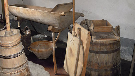 Fotoaufnahme von Vorratsgefäßen und Küchengerrät aus Holz in einer Kammer, deren Tapete mit figuerlichen Verzierungen bemalt wurde