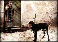Vor einer Haustüre steht ein älterer Herr in Arbeitskleidung, gestützt auf einen Stock. Im Vordergrund steht ein Hund, am Haus angekettet.