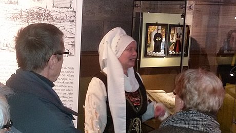 Fotoaufnahme einer Erlebnisführung durch die Spitalkirche. Im Fokus steht eine als Argula von Grumbach kostümierte Museumsführerin. Vor ihr stehen einige Besucher, die ihren Erzählungen lauschen.  Hinter ihr befinden sich eine Infotafel und eine Vitrine mit zwei Bildern. 