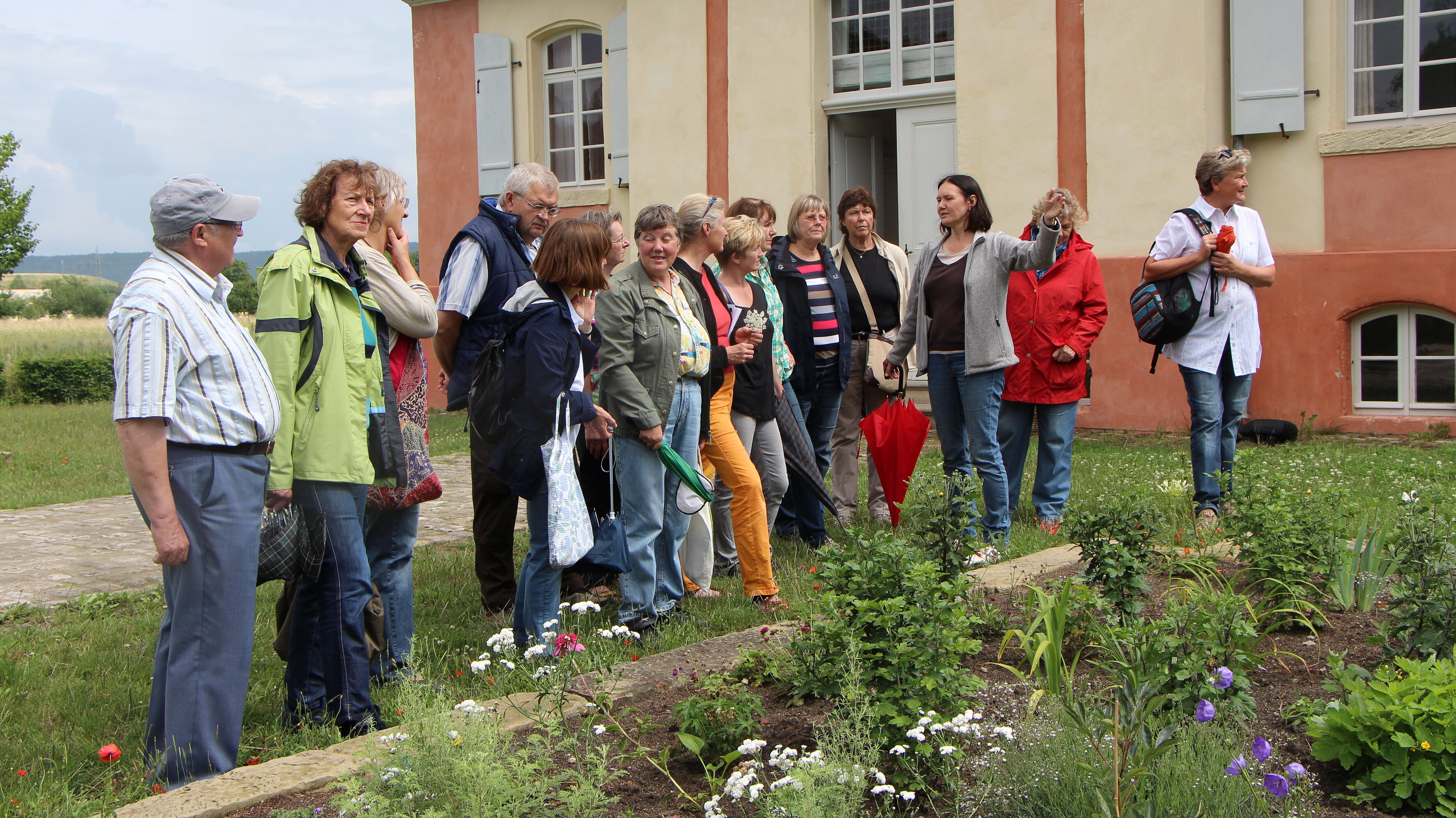 Fotoaufnahme einer Führung im Garten des Jagdschlösschens aus Eyerlohe. Die Gruppe steht vor dem angelegten Blumenbeet. Es blühen unter anderen Staudenpfingstrosen, Mohn und Clematis. Hinter der Gruppe befindet sich das Schlösschen mit der geöffneten Tür. 