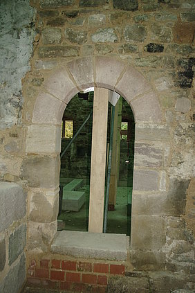 Fotoaufnahme einer fertiggestellten Tür im Badhaus. Das Foto wurde im Flur aufgenommen. Gezeigt wird der runde Torbogen aus Sandsteinquadern, eingebettet in das Steinmauerwerk. Im Raum dahinter sind aufgestellte Holzbalken und Metallstreben erkennbar. 