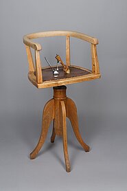 Der Stuhl ist aus Holz. Er ist drehbar. Er hat vier Stuhlbeine in geschwungener Form. Sie sind an einer Säule befestigt. Auf der Säule ist die Sitzfläche. An der Sitzfläche ist eine niedrige Lehne. Sie ist auch eine Armlehne. Auf dem Stuhl liegt ein Gerät zum Haare schneiden.