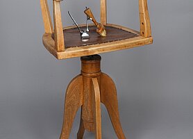 Der Stuhl ist aus Holz. Er ist drehbar. Er hat vier Stuhlbeine in geschwungener Form. Sie sind an einer Säule befestigt. Auf der Säule ist die Sitzfläche. An der Sitzfläche ist eine niedrige Lehne. Sie ist auch eine Armlehne. Auf dem Stuhl liegt ein Gerät zum Haare schneiden.