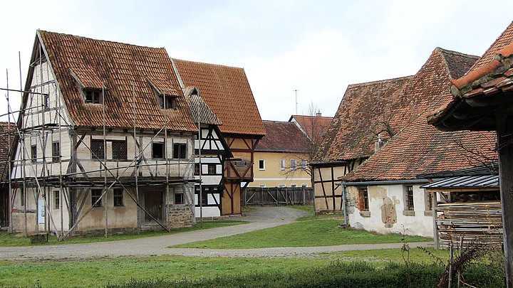 Fotoaufnahme der Gasse mittelalterlicher Fachwerkhäuser in der Baugruppe Stadt. An einem Weg befinden sich auf beiden Seiten verschiedene Fachwerkhäuser. Vorne links steht zum Beispiel das Handwerkerhaus aus Bad Windsheim, das von einem Holzgerüst umgeben ist.  