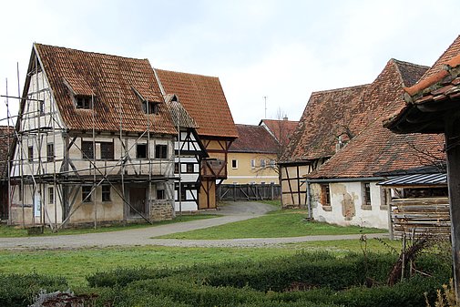 Fotoaufnahme der Gasse mittelalterlicher Fachwerkhäuser in der Baugruppe Stadt. An einem Weg befinden sich auf beiden Seiten verschiedene Fachwerkhäuser. Vorne links steht zum Beispiel das Handwerkerhaus aus Bad Windsheim, das von einem Holzgerüst umgeben ist.  