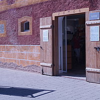 Fotoaufnahme des Eingangsbereiches der Kasse, die sich im Korbhaus aus Knittelsbach befindet. In den Rahmen der geöffneten Holztür wurden die Jahreszahl „2006“, sowie das Fränkische Wappen eingeprägt. An der Tür hängen mehrere Schilder. 