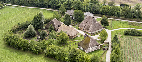 Fotoaufnahme der Baugruppe Mittelalter von einem erhöhten Standpunkt. Die Häuser stehen im Mittelpunkt des Bildes, außenherum befinden sich Wiesen, Felder und Bäume. Auf den Wegen sind einige Besucher erkennbar. 