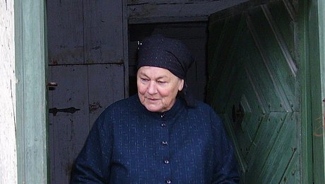 Fotoaufnahme der Führung „Zu Besuch bei Maria Dörfler“. In der geöffneten Tür des Bauernhauses aus Oberzettlitz steht eine als Maria Dörfler kostümierte Museumsführerin. Sie trägt einen zugeknöpften Kittel und ein Kopftuch. 