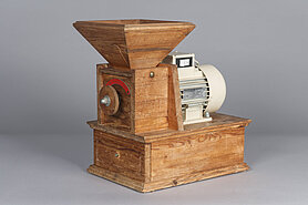 Die Mühle ist aus Holz. Unten ist ein Kasten mit Schublade. Darauf ist ein kleinerer Kasten mit Drehrad. Oben auf dem kleineren Kasten ist ein Trichter. Neben dem kleineren Kasten ist ein Motor.