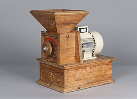 Die Mühle ist aus Holz. Unten ist ein Kasten mit Schublade. Darauf ist ein kleinerer Kasten mit Drehrad. Oben auf dem kleineren Kasten ist ein Trichter. Neben dem kleineren Kasten ist ein Motor.
