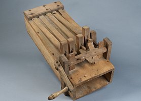 Die Ratsche ist ein Holzkasten. Auf dem Holzkasten sind Holzleisten und eine Kurbel. Die Kurbel zieht an den Holzleisten. Die Holzleisten schlagen auf den Kasten. Es ist ein lautes Knallen zu hören.