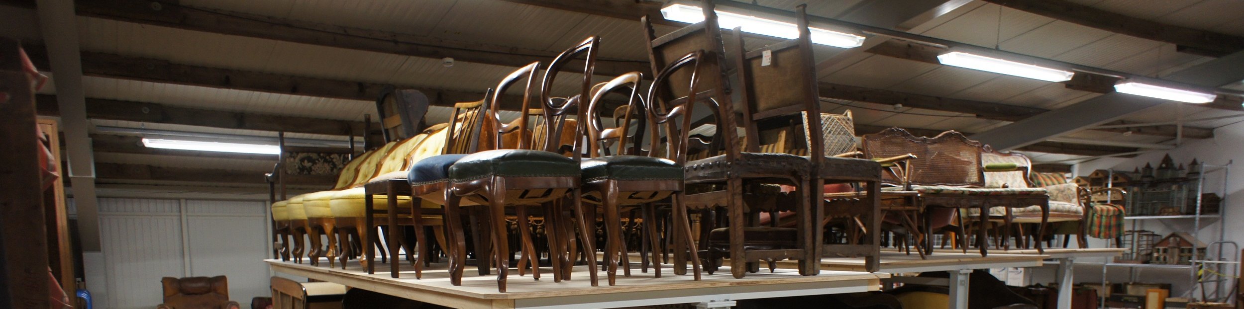 Fotoaufnahme vom Inneren des Möbeldepots. Auf den Platten von mehreren übergroßen Tischen befinden sich etliche Stühle. Unter den Tischen stehen Sessel und Sofas auf Transportroller. An den Wänden im Hintergrund sind Regale mit Objekten erkennbar. 