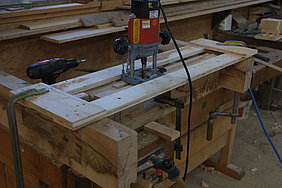 Ein Kopfholz ist in die Schraubzwinge der Werkbank eingespannt. Mit einem Werkzeug wird auf der Längsseite des Kopfholzes die Verbindung für die Schalbretter eingearbeitet.
