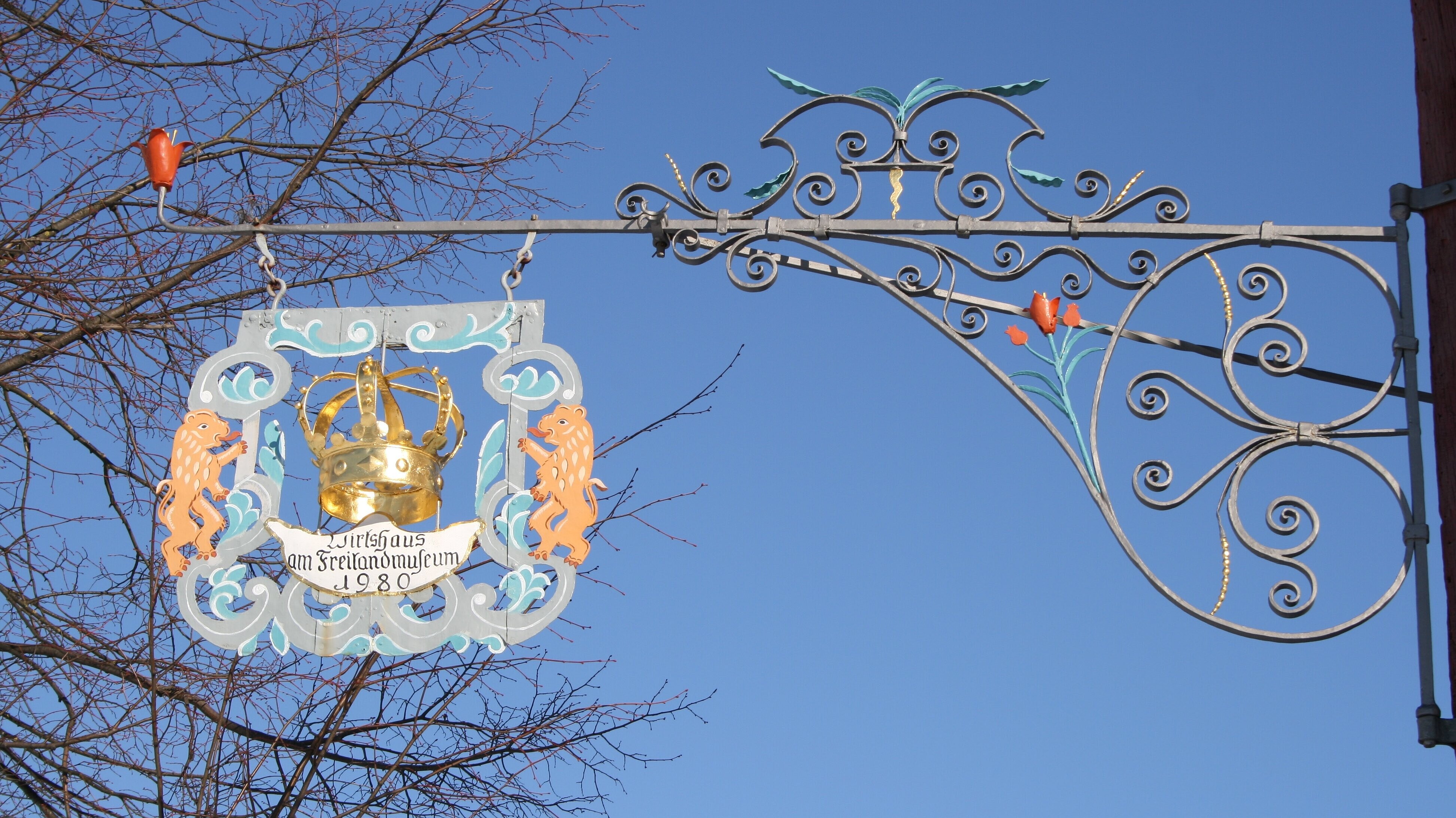 FFM_Ausleger_Oberampfrach Fotoaufnahme des Auslegers vom Gasthaus zur Krone aus Oberampfrach. Das Nasenschild ist im rechten Winkel an der Mauer befestigt. In der Mitte befindet sich eine Krone, darunter steht „Wirtshaus am Freilandmuseum 1960“. An den Seiten wird das Schild von je einem Löwen gehalten.