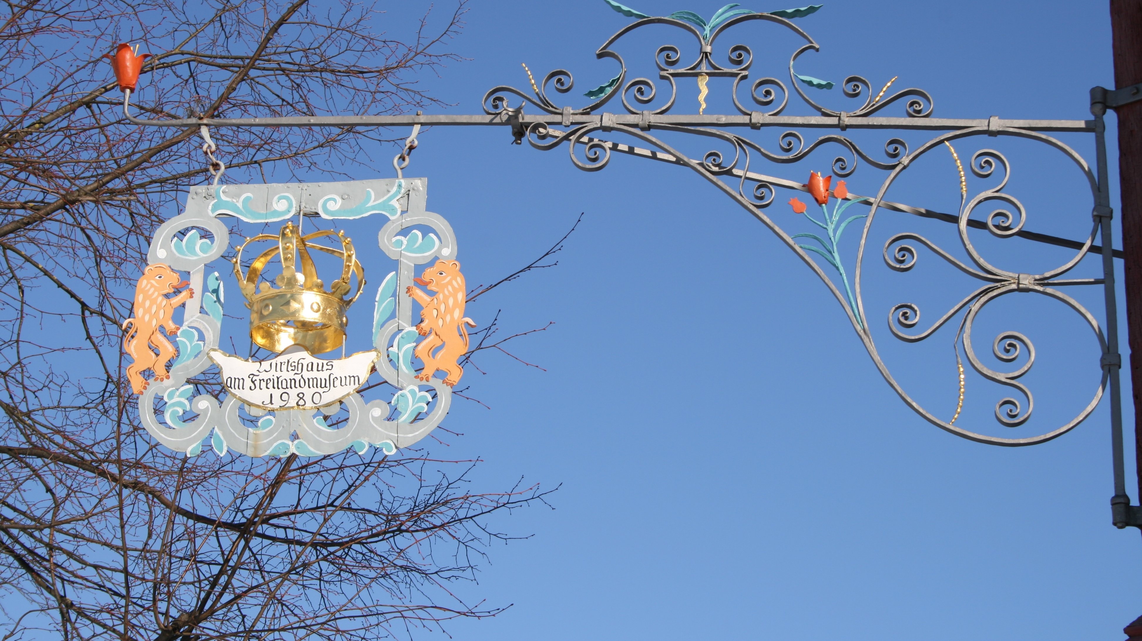 Fotoaufnahme des Auslegers vom Gasthaus zur Krone aus Oberampfrach. Das Nasenschild ist im rechten Winkel an der Mauer befestigt. In der Mitte befindet sich eine Krone, darunter steht „Wirtshaus am Freilandmuseum 1960“. An den Seiten wird das Schild von je einem Löwen gehalten.