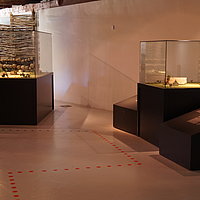 Fotoaufnahme der Ausstellung des Archäologiemuseums, welches sich in der Schafscheune aus Virnsberg befindet. Gezeigt werden zwei Vitrinen, in denen sich Modele von Siedlungen bzw. Häusern befinden. Auf dem Boden ist ein Leitsystem durch die Ausstellung erkennbar. 