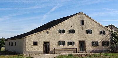 Fotoaufnahme des Bauernhauses aus Reichersdorf am aktuellen Standort. Das breitgelagerte eingeschossige Jurahaus hat zwei Eingänge an der Giebelseite. Links ist die Stalltür, rechts die Haustür aus Holz. Die Fenster auf der Wohnseite haben geöffnete Läden. 