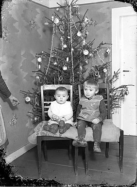 Die zwei Kinder sitzen vor einem Weihnachtsbaum. Der Baum ist geschmückt. Die Kinder tragen Schürzen. Eine Schürze hat einen aufgestickten Anker. Die andere Schürze ist weiß.