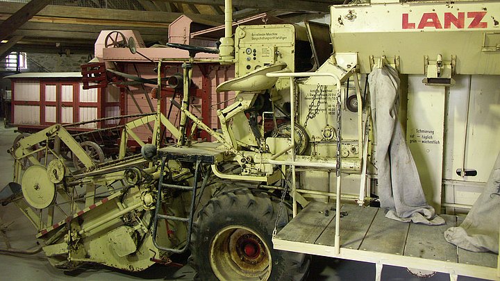 Fotoaufnahme der Dauerausstellung zur Mechanisierung der Landwirtschaft in Franken in der Schafscheuer aus Weiltingen. Im Vordergrund steht ein Lanz-Mähdrescher, dahinter eine Elektro-Dreschmaschine. An der Decke ist der offene Dachstuhl erkennbar. 