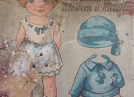 Auf dem DEckel ist ein Kind abgebildet. Es trägt Unterwäsche. Daneben sind eine Mütze und ein Kleid. Beides ist blau.