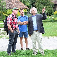 Fotoaufnahme zweier Besucher mit einem Museumsführer. Die drei Männer betrachten etwas außerhalb des Bildes. Einer steht auf dem Kiesweg, die anderen beiden im Gras. Im Hintergrund befinden sich eine Bierzeltgarnitur sowie Büsche, Blumen und Gebäude. 