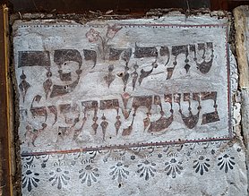 Das Gefach war Teil einer Fachwerkwand. Auf dem Gefach ist ein kurzer Text. Der Text ist in hebräischen Buchstaben geschrieben. Um den Text sind Verizerungen.